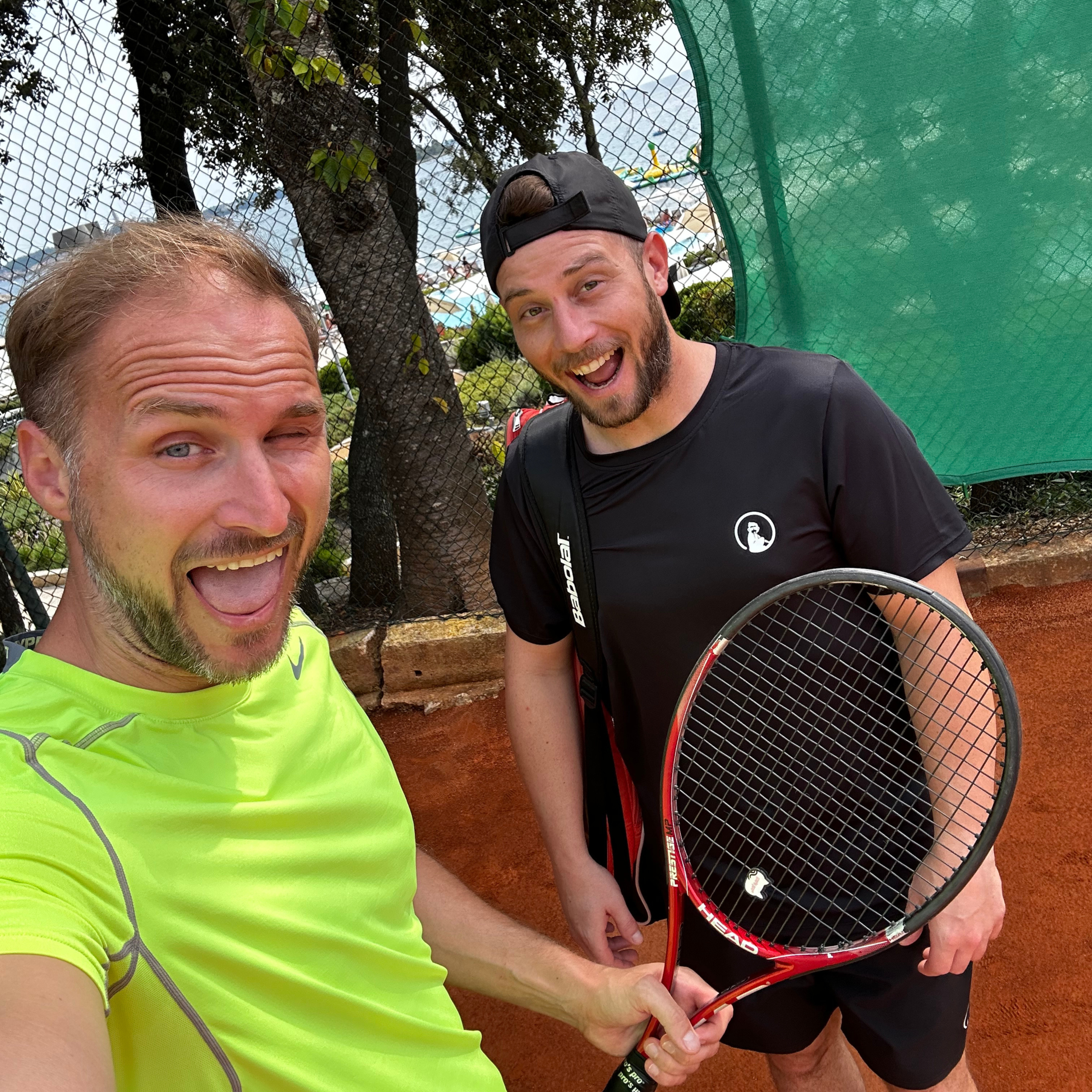 Zwei Kollegen lachen auf einem Sandtennisplatz mit Tennisschlägern.